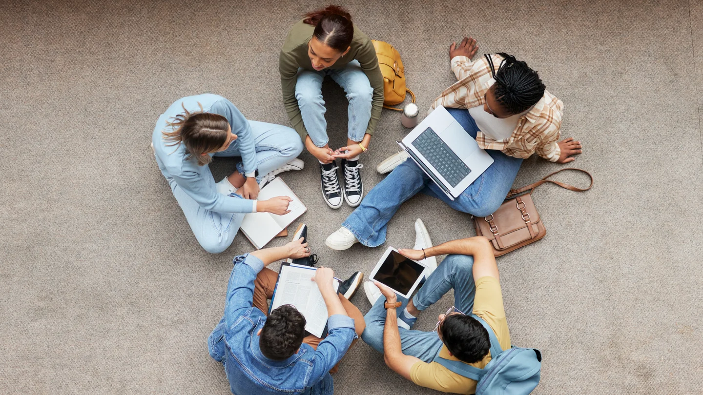 Junge Studierende mit Arbeitsmaterialien sitzen auf dem Fußboden im Kreis.