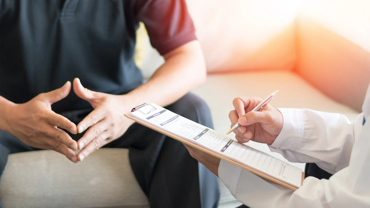 Syphilis: Ein Mann sitzt auf einer Couch, seine Hände Formen eine Raute. Schräg vor ihm sitzt ein Arzt, der ein Klemmbrett und einen Kugelschreiber in den Händen hält.