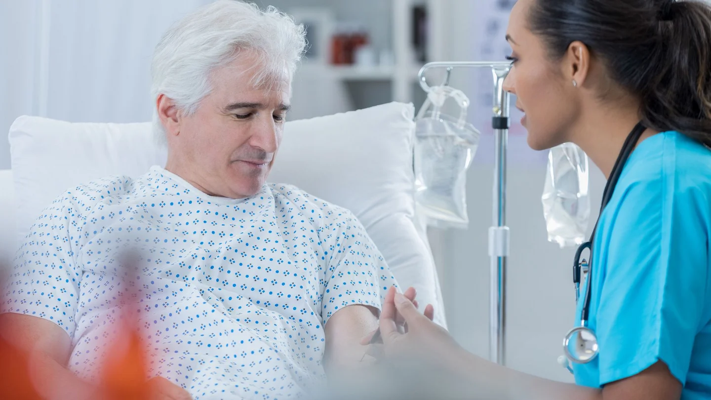 Prostatakrebs: Ein älterer Mann sitzt in einem Krankenhausbett. Er schaut nach unten, ihm gegenüber sitzt eine Krankenschwester. Sie schaut ihn an und entnimmt ihm Blut.