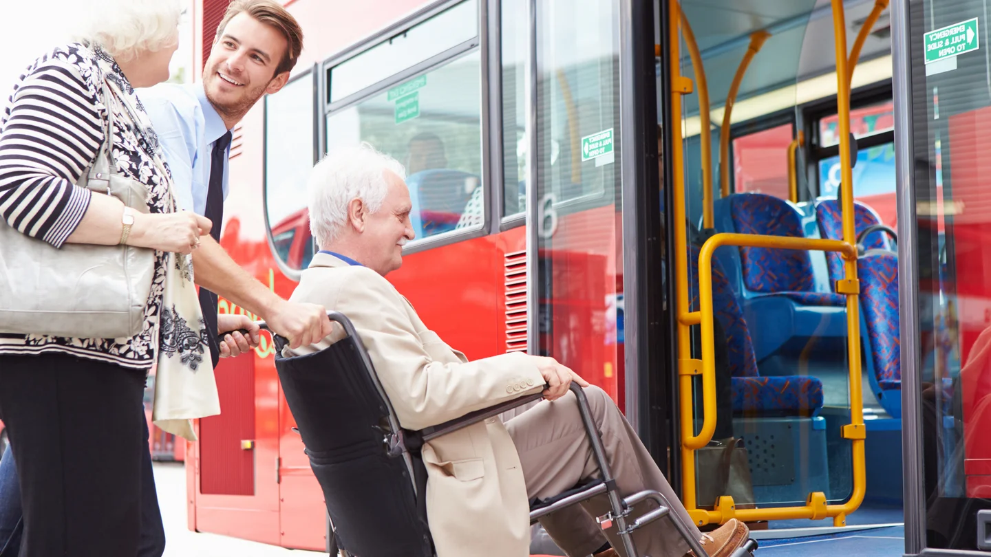 Ein junger Busfahrer schiebt einen älteren Herrn im Rollstuhl über eine Rampe in den Bus.
