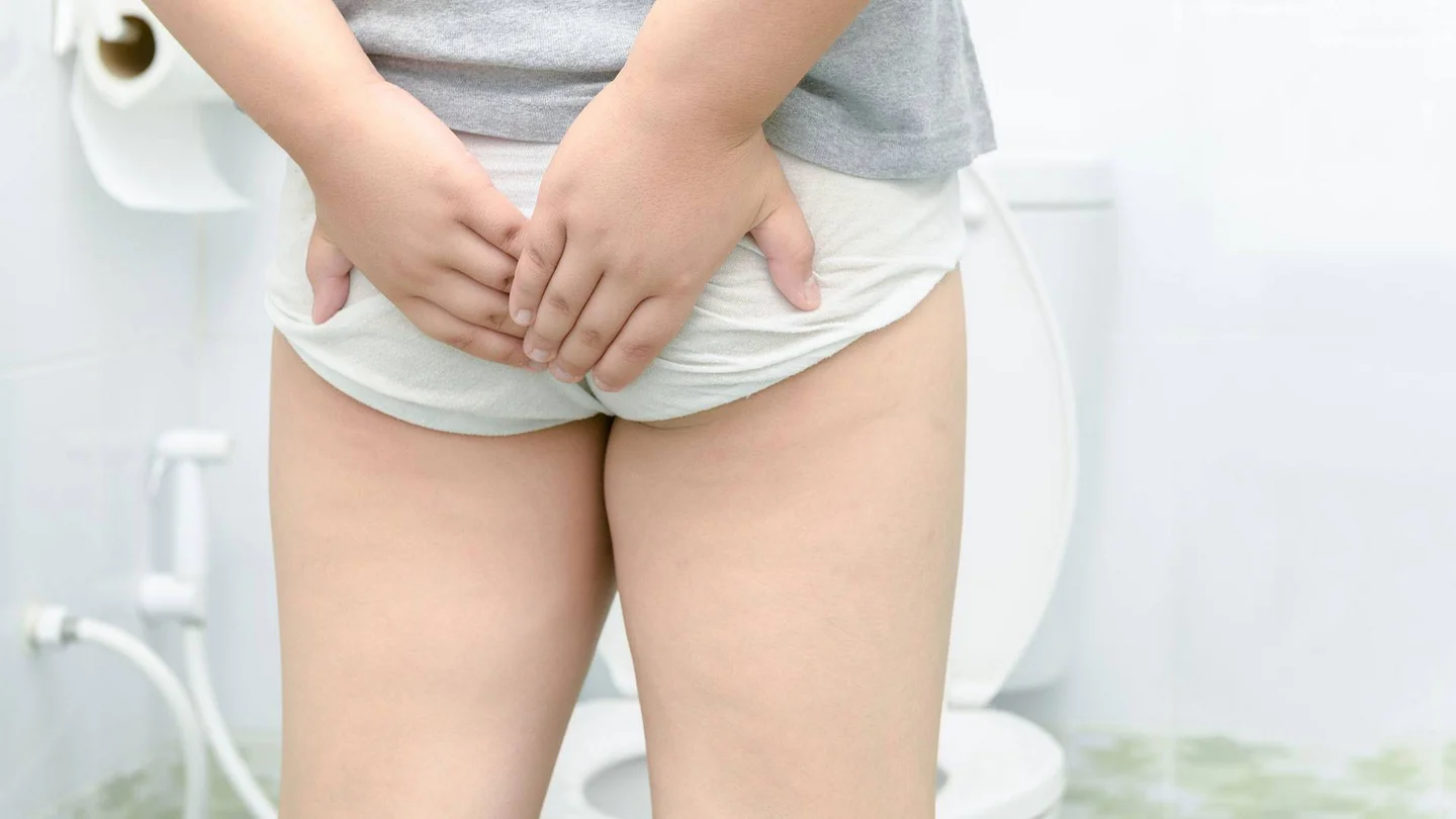 Kind, mit Unterhose bekleidet, fasst sich an das Gesäß. Im Hintergrund eine Toilette.