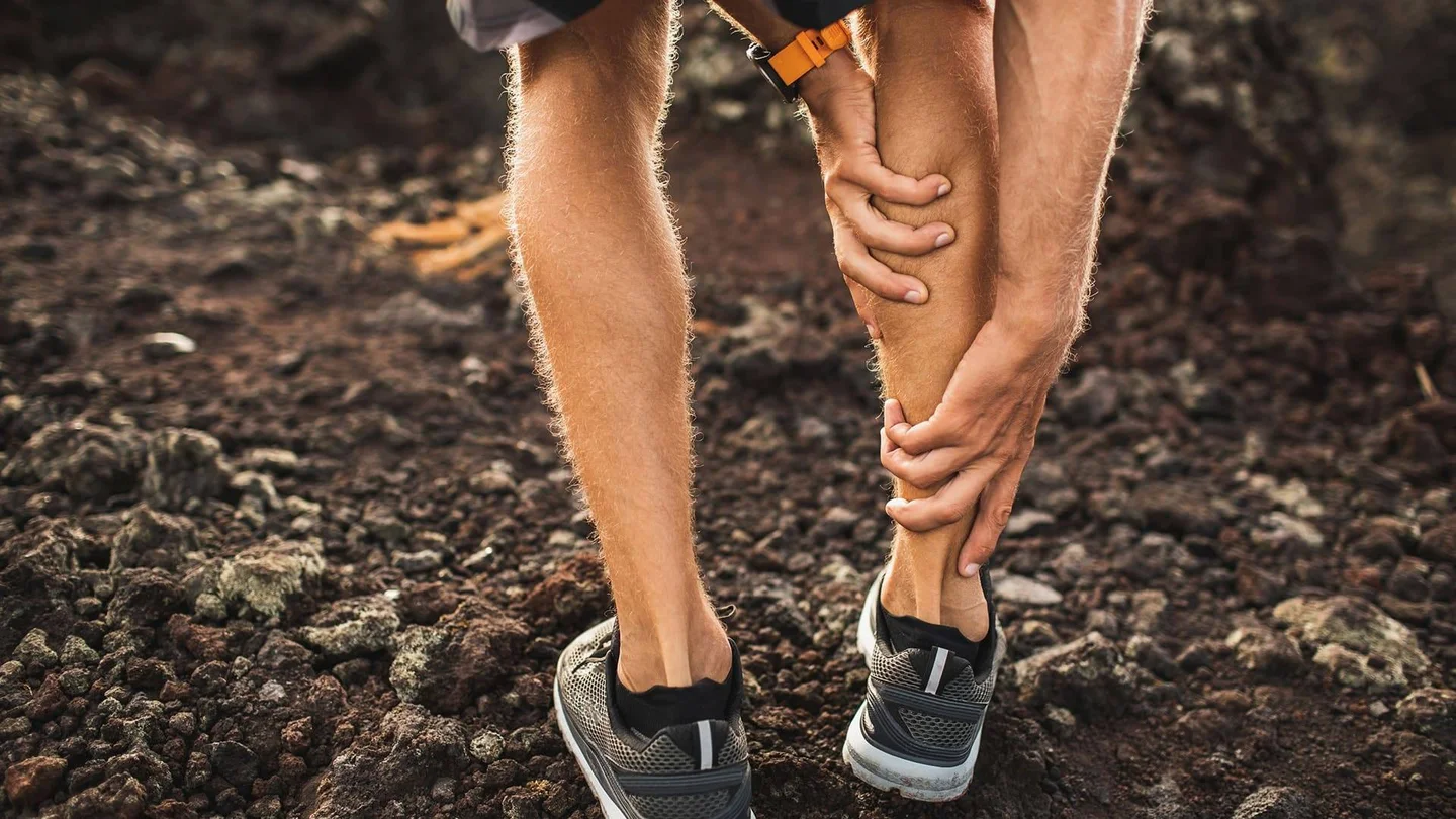 Eine Person in Sportkleidung bückt sich und fasst sich an die vermutlich schmerzende Wade.