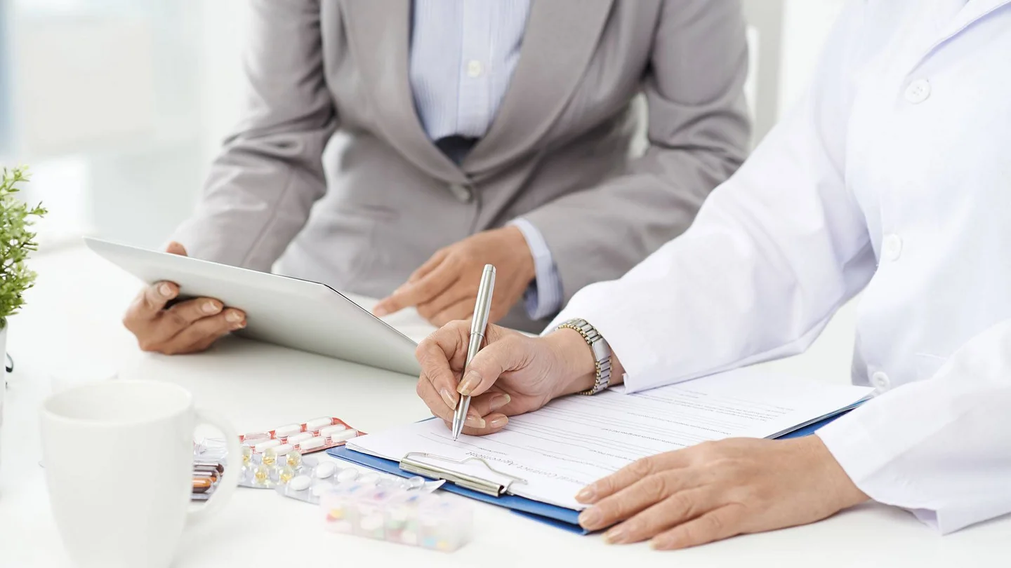 Eine Person im weißen Kittel füllt ein Dokument aus. Daneben sitzt eine Person, die auf ein Tablet zeigt.