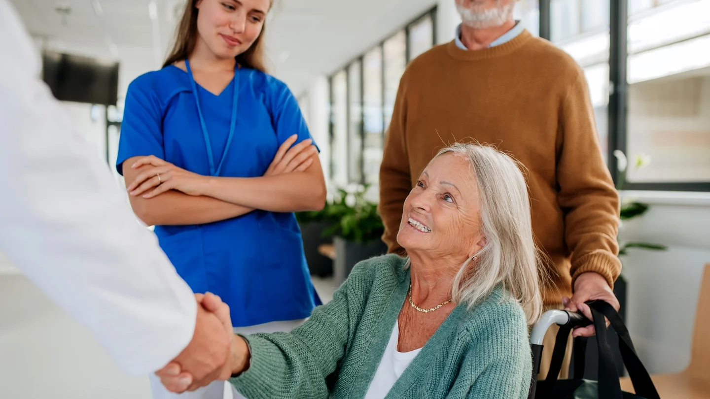Eine &auml;ltere Frau sitzt in einem Rollstuhl in einem hellen Krankenhausflur. Sie l&auml;chelt und gibt einer &Auml;rztin oder einem Arzt die Hand. Im Hintergrund stehen eine Krankenpflegerin und ein &auml;lterer Mann, der den Rollstuhl schiebt und eine Tasche tr&auml;gt.