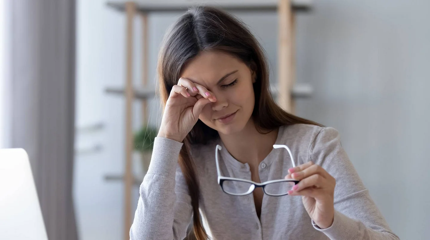 Frau hat die Brille abgenommen und reibt sich die Augen. Neben ihr ein Laptop.