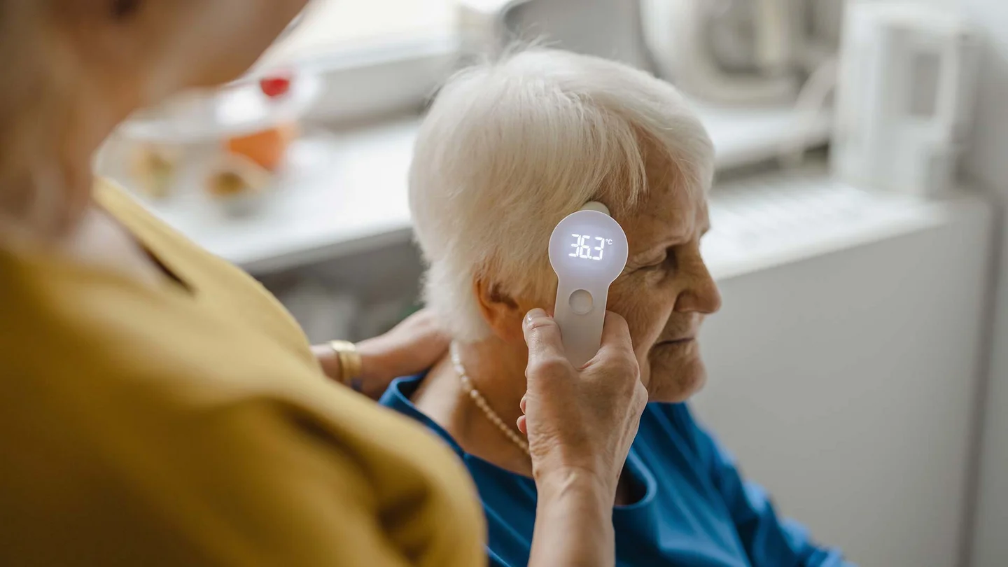 Pflegekraft überprüft Temperatur einer Seniorin.
