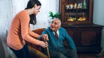 Ein alter Herr möchte aus seinem Wohnzimmersessel aufstehen, er hat einen Gehstock zur Hilfe. Eine junge Frau hilft ihm bei dem Aufstehen und greift ihm unter den Arm als Stütze.