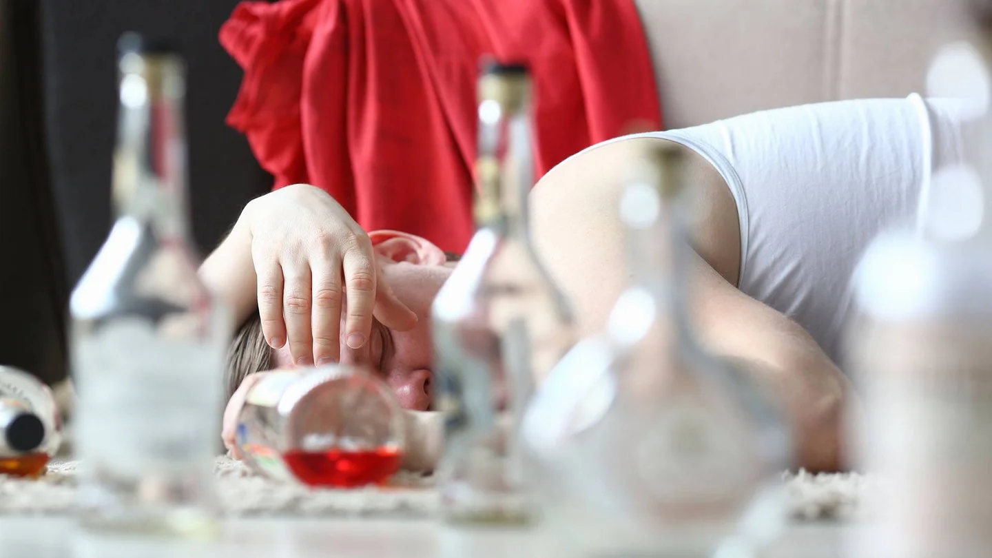 Auf einem Tisch sind verschwommen Flaschen und Gläser zu sehen. Eine Person liegt daneben und schläft.
