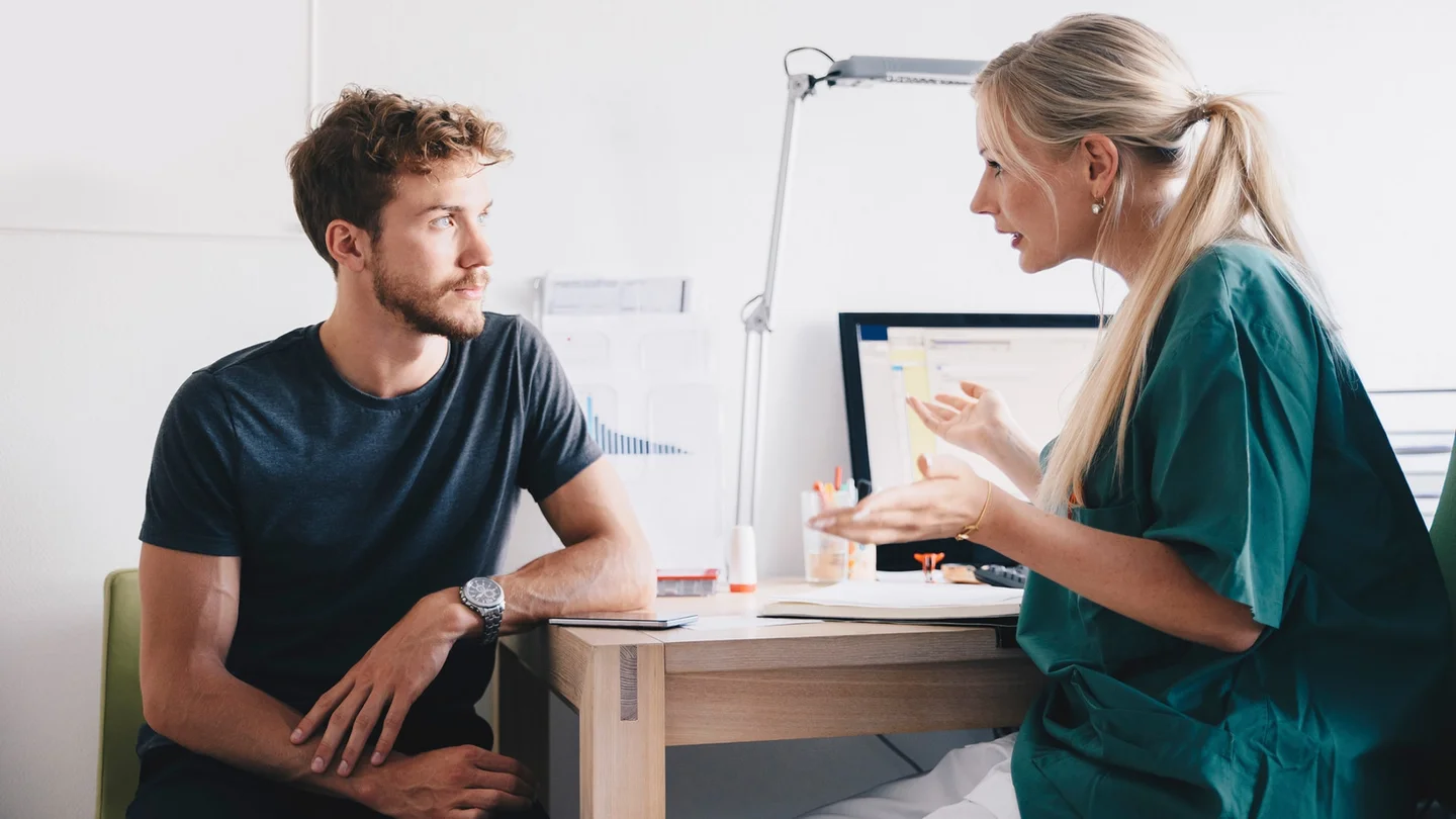 Hodenkrebs: Ein Mann sitzt in einem Behandlungszimmer. Sein Blick ist auf eine Ärztin gerichtet, die ihm gegenüber sitzt und mit ihm spricht.