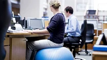 Gesundheit am Arbeitsplatz: Eine Frau sitzt auf einem Gymnastikball am Schreibtisch und schaut nach rechts zu einem Display.