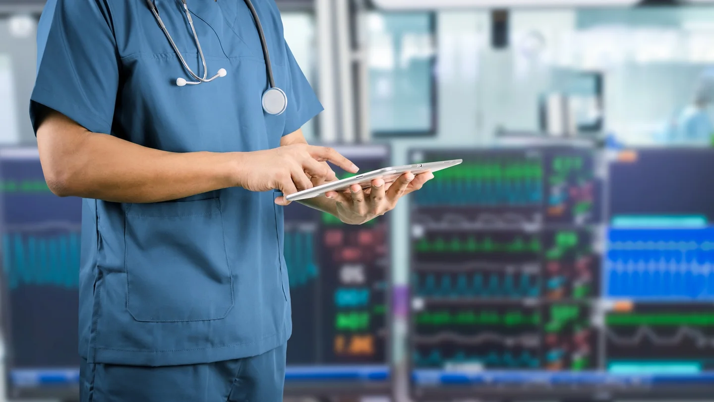 Eine medizinische Fachkraft bedient ein Tablet, im Hintergrund sind Monitore zur Kreislauf&uuml;berwachung sichtbar.